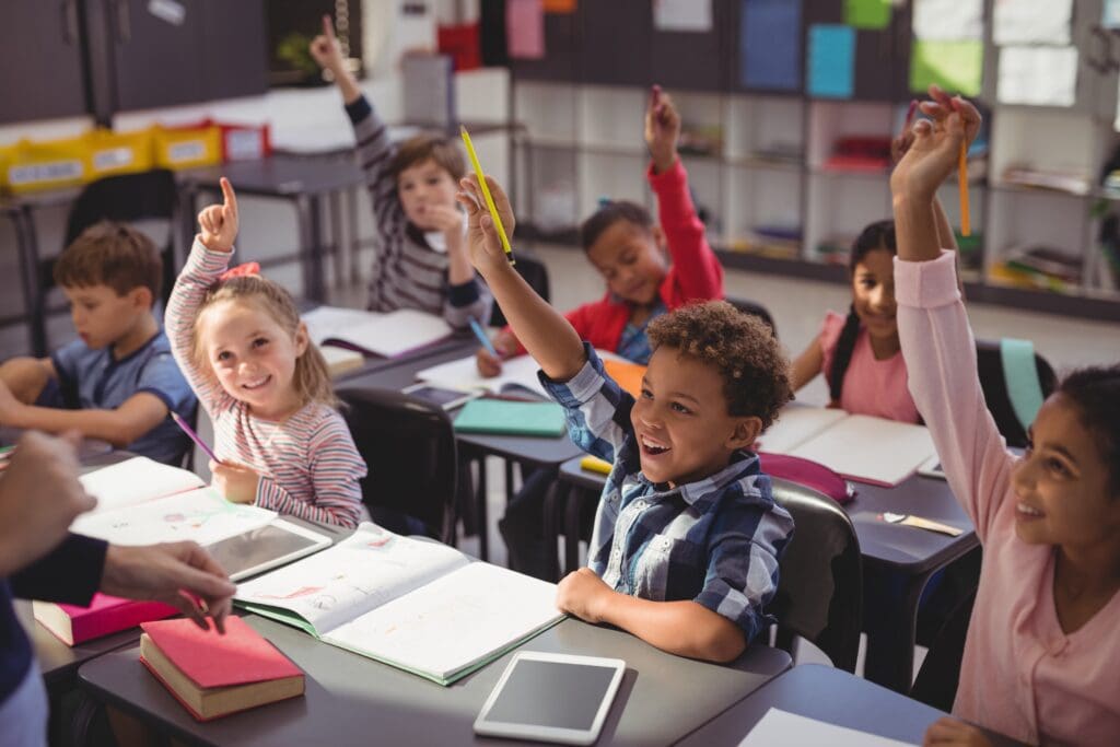 A classroom of young learners, all smiling with their hands raised.