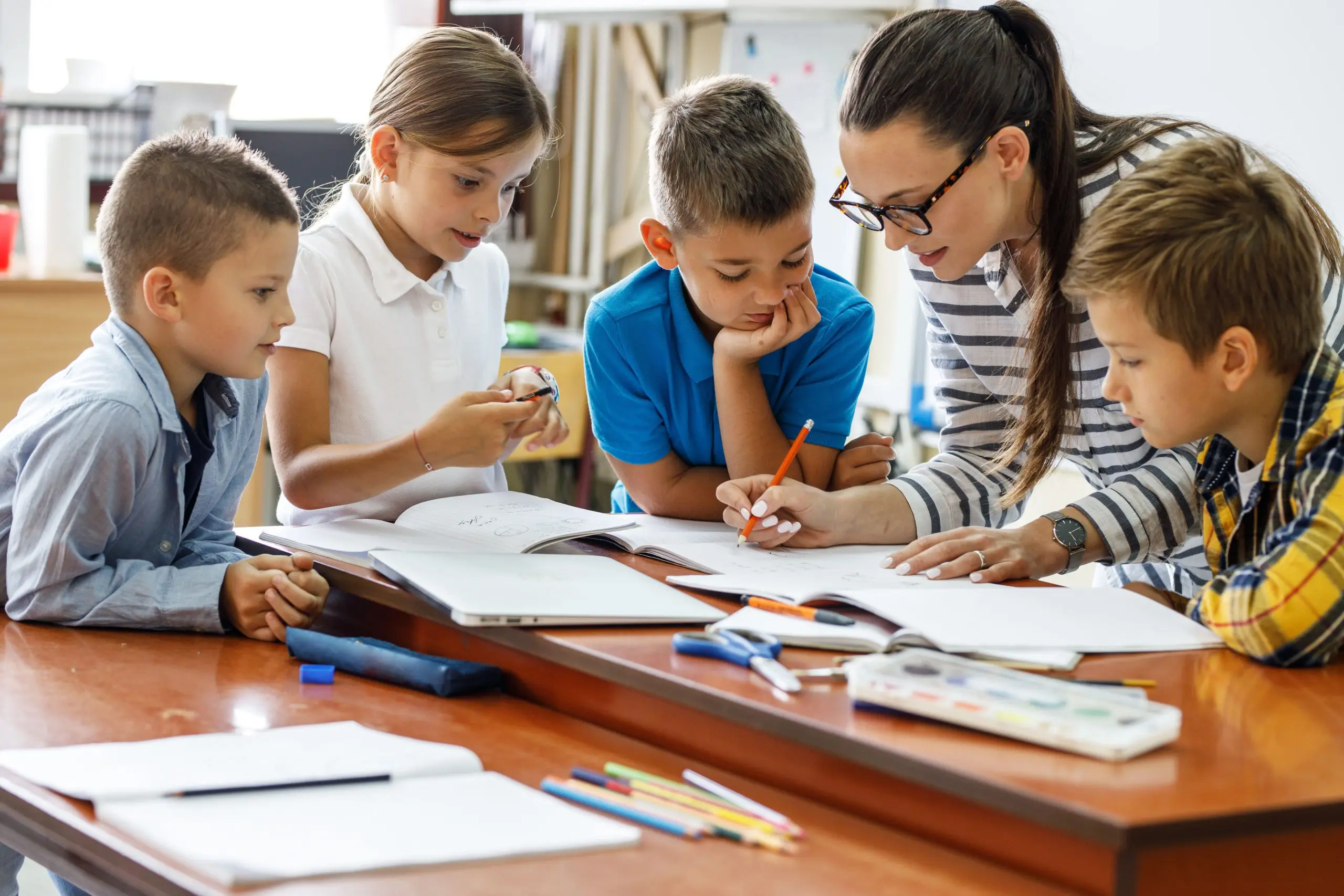 A teacher writes in a notebook on a desk while school children gather around and watch her.