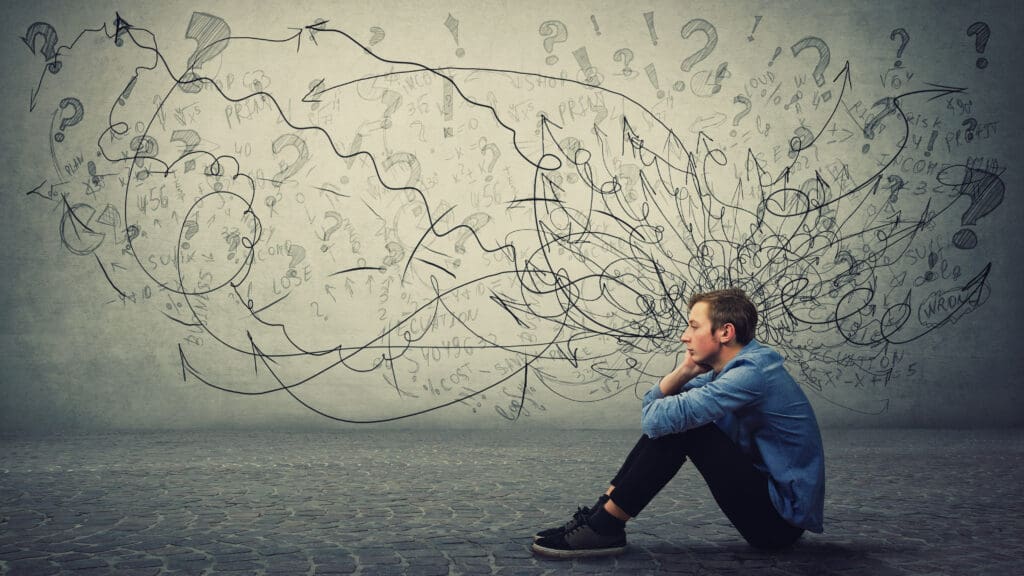 A teenager sitting down and random scribbles, arrows, and question marks are on the wall behind him, seemingly coming out of his head.